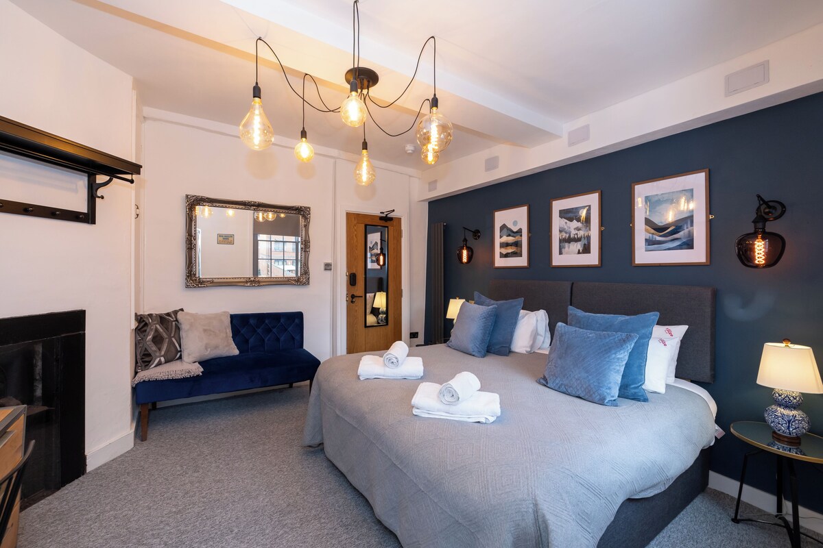 A hotel room with a large bed, a blue sofa, a fireplace, and a wall with framed pictures.