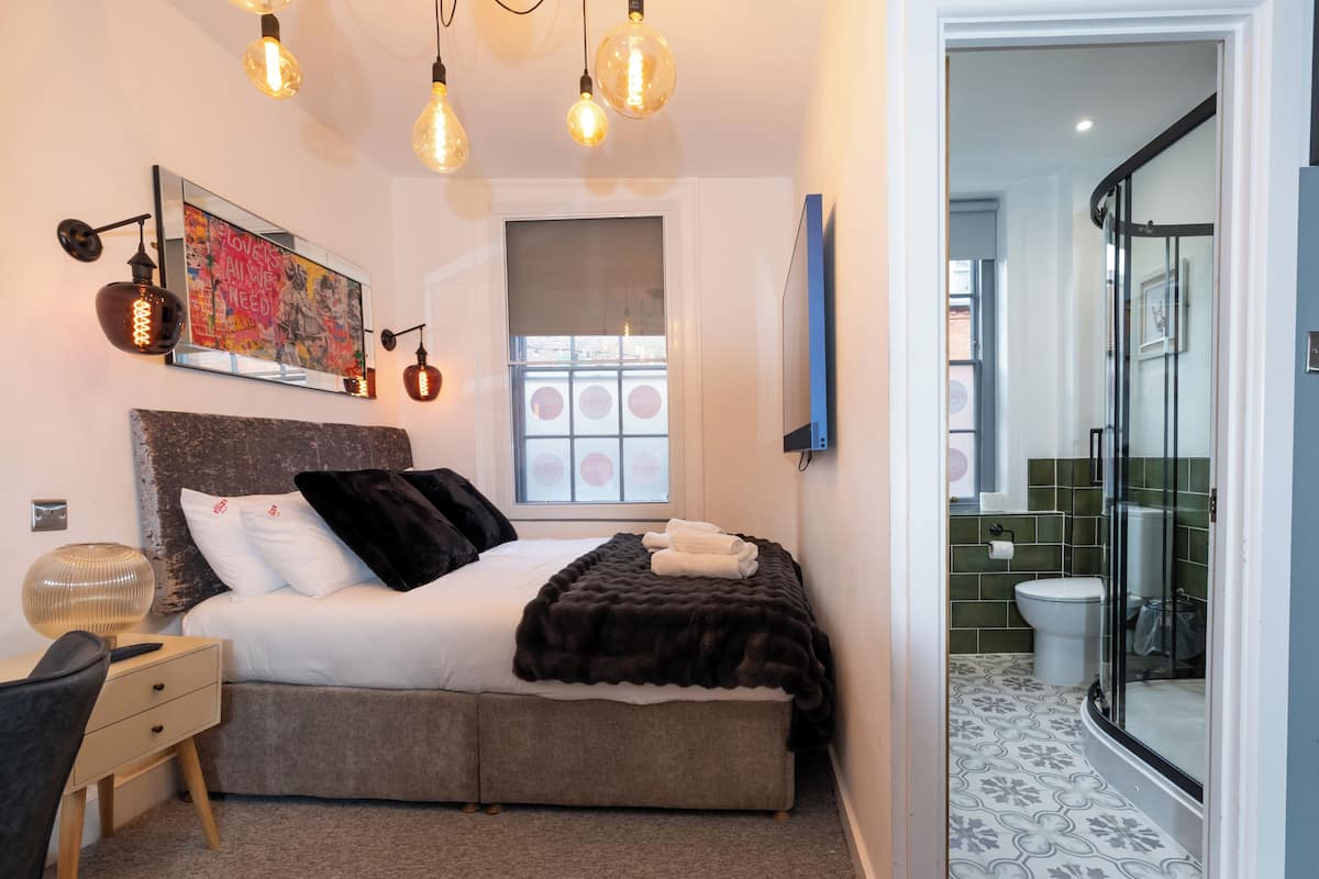 A bedroom with a bed, bedside table, lamp, artwork, and a window.