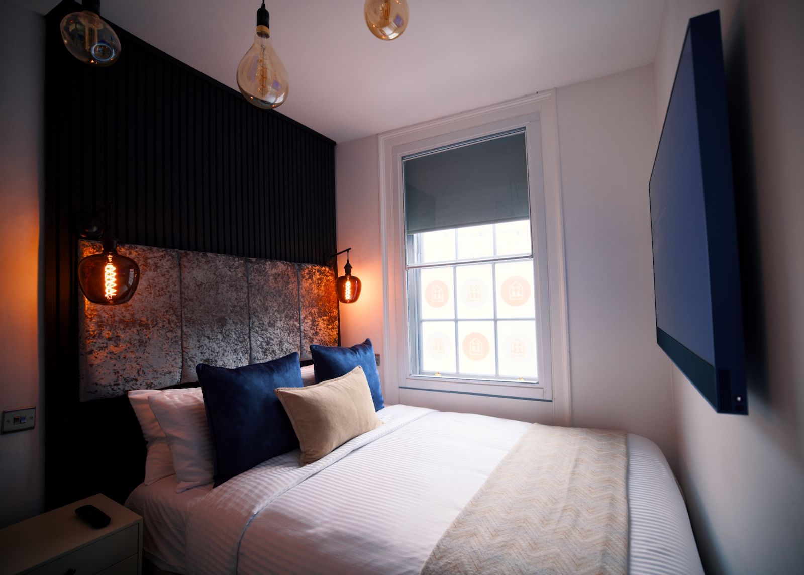 A bedroom with a bed, pillows, a nightstand, a wall-mounted TV, and two hanging light fixtures.