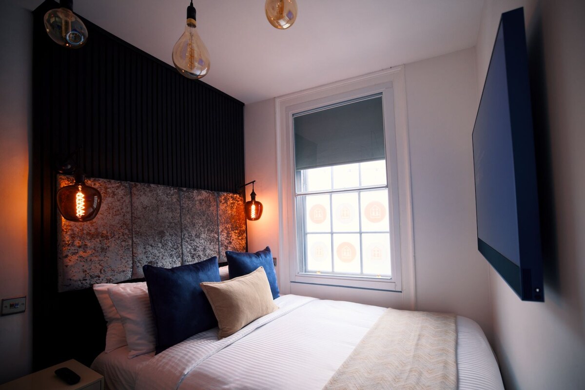 A bedroom with a bed, pillows, a nightstand, a wall-mounted TV, and two hanging light fixtures.