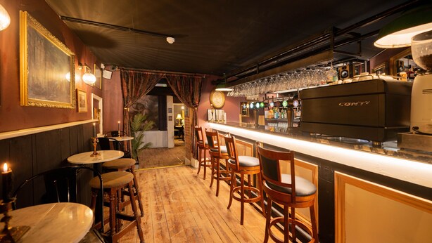 A bar area with wooden bar stools, a counter with a coffee machine, and a wall with framed pictures. - City Rooms by Hiive (Worcester)