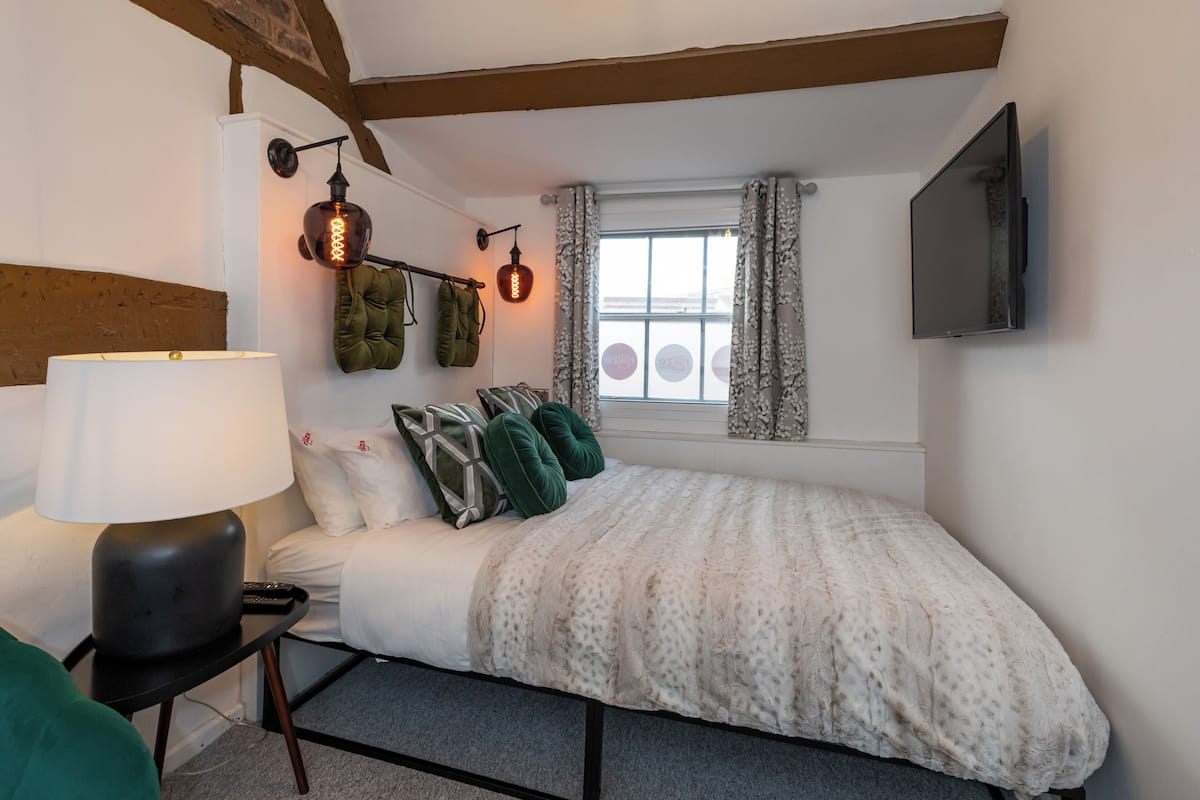 A bedroom with a bed, a lamp, a TV, a window with curtains, and a brick wall.