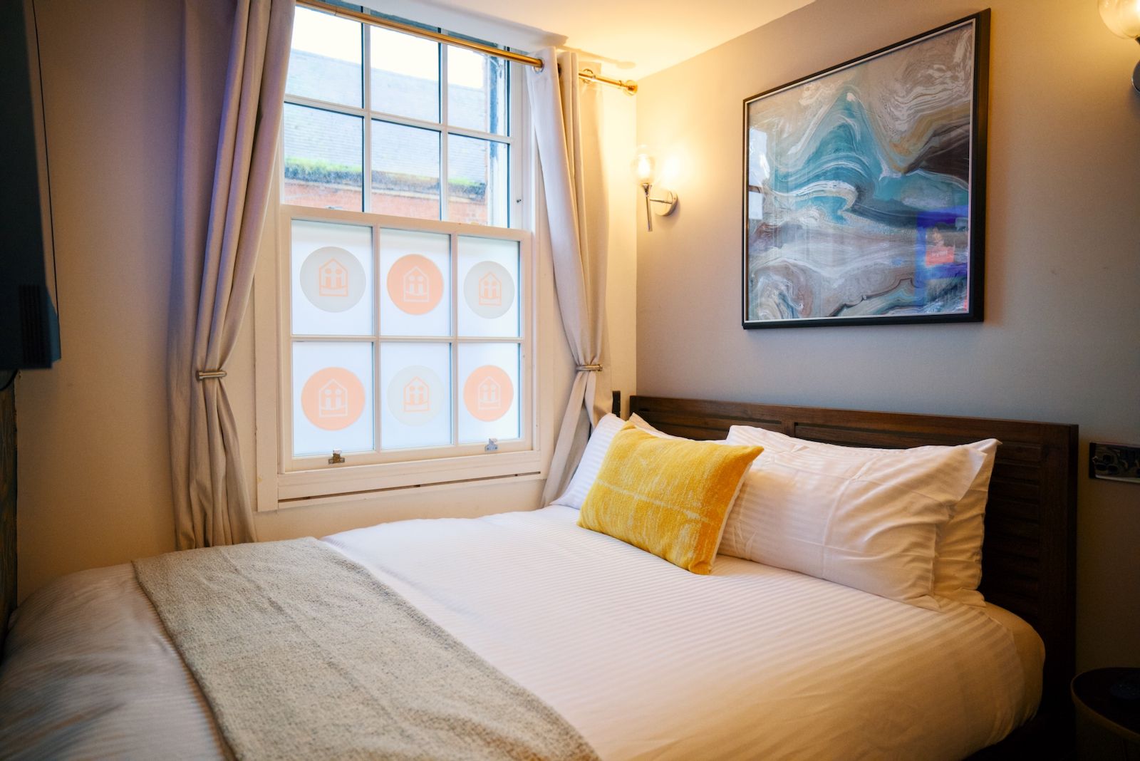 Una cama bien hecha con un cojín blanco y amarillo, una ventana con cortinas y un cuadro abstracto colgado en la pared.