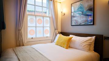 Una cama bien hecha con un cojín blanco y amarillo, una ventana con cortinas y un cuadro abstracto colgado en la pared.