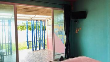 Una habitación con una cama, un televisor fijado a la pared y un ventanal corredero que da a un espacio exterior con cercado azul y vegetación.