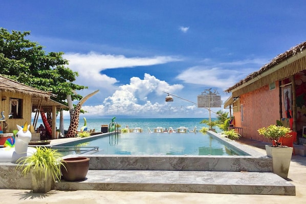 A pool area with a view of the ocean, lounge chairs, and a thatched-roof building.