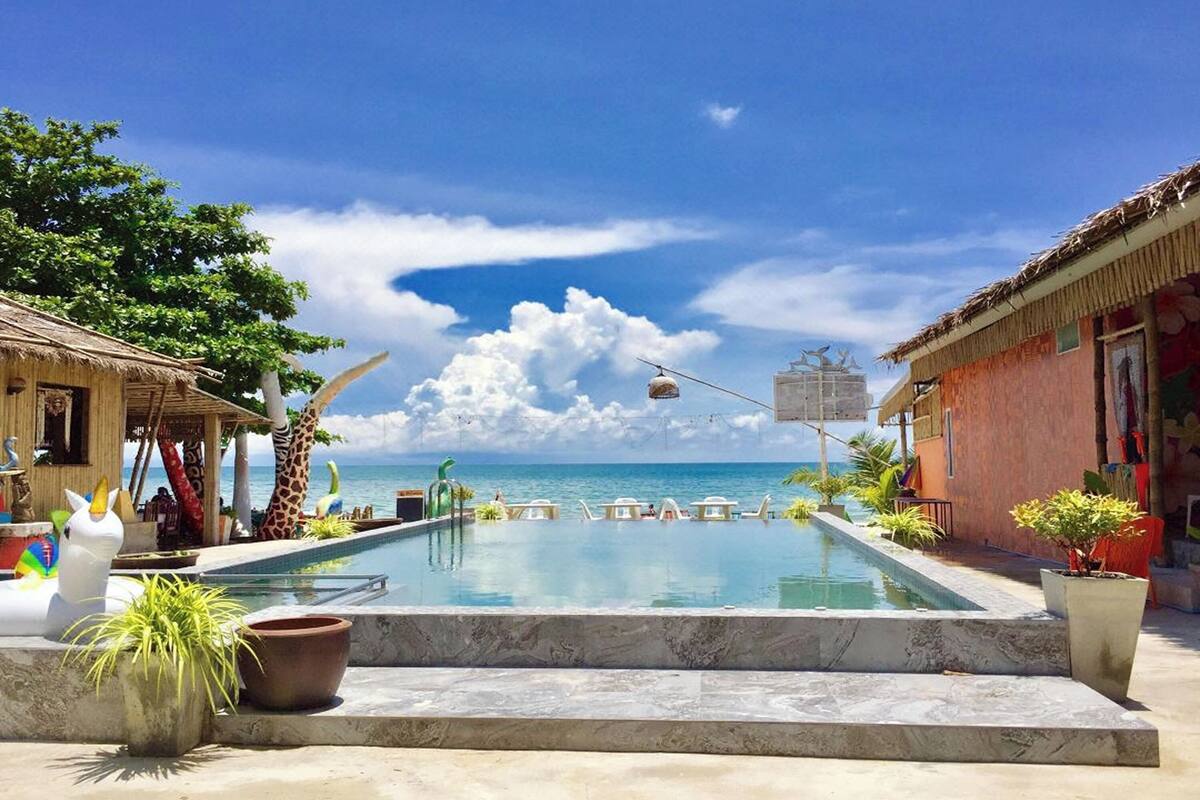Zona de piscina con vista al océano, sillas de descanso y un edificio con techo de paja.