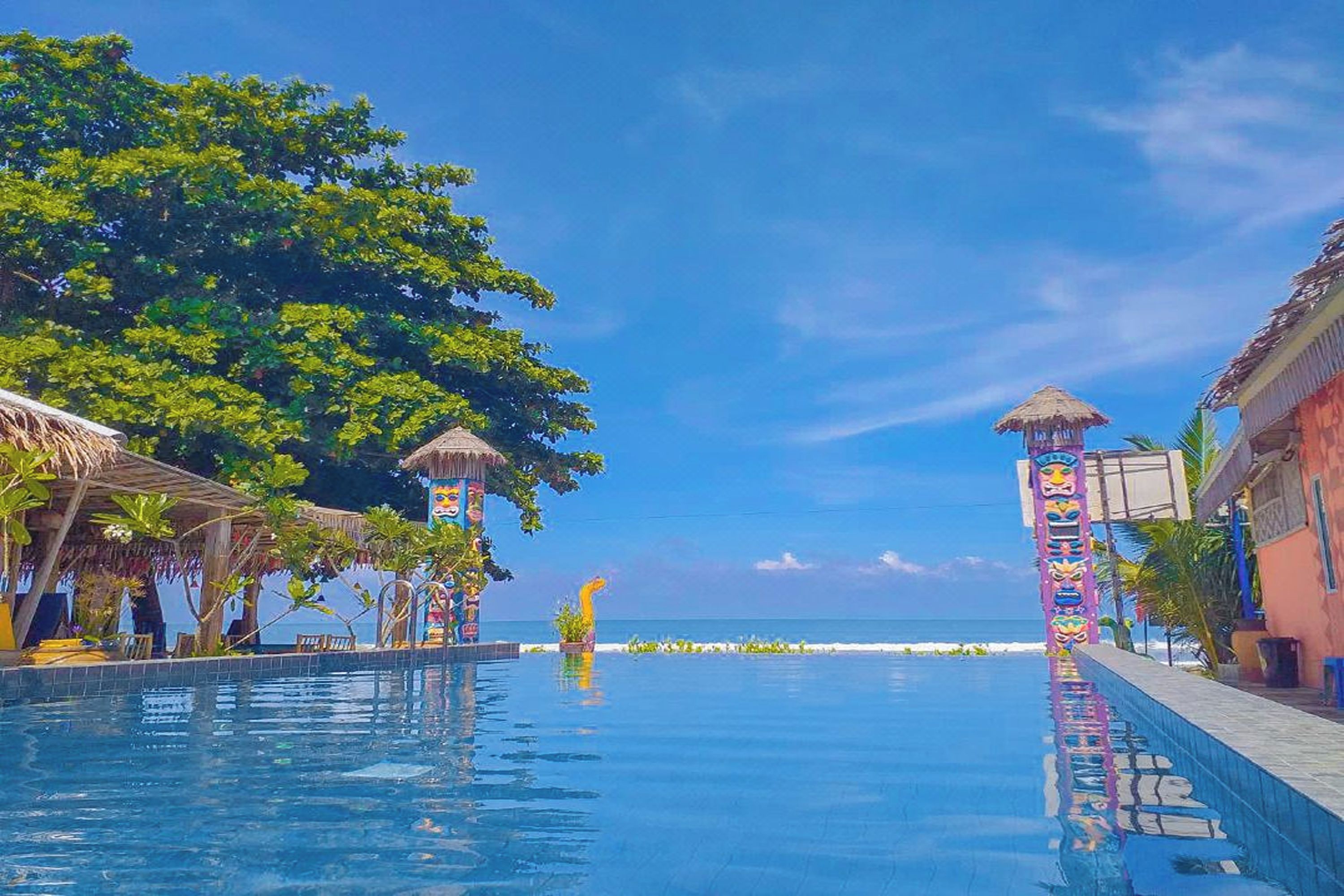 Una piscina con vistas claras al océano, rodeada de construcciones con techo de paja y una decoración vertical colorida.