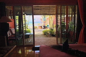 Una habitación con una cama, una mesa y una silla. Hay una puerta corrediza de vidrio que da a un espacio exterior con plantas y vistas al océano.