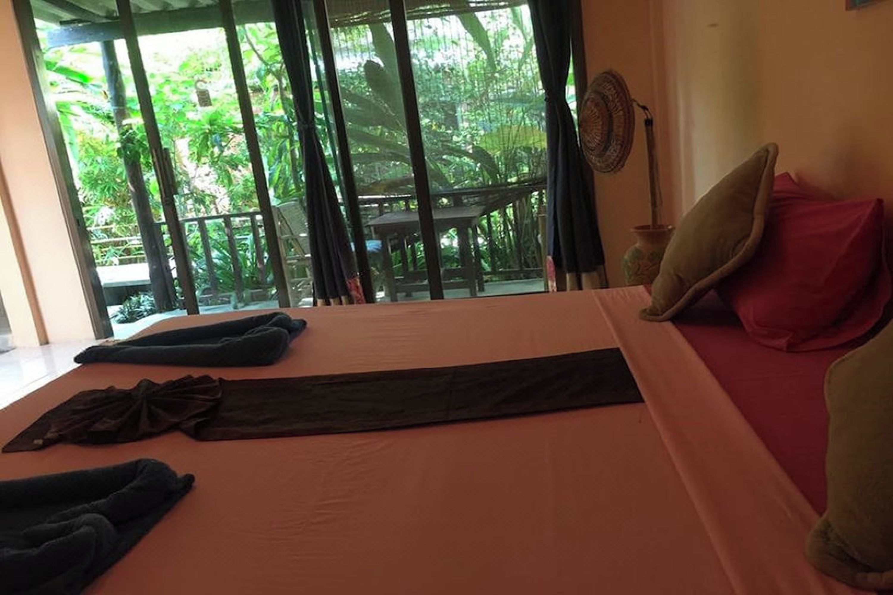 A room with a bed, a window with a view of greenery, and a fan on the wall.