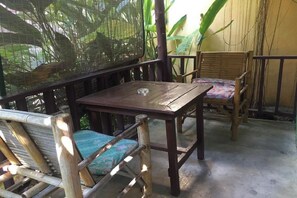 Una mesa de madera y dos sillas con asientos acolchados, ubicadas en un balcón con mosquitero y plantas al fondo.