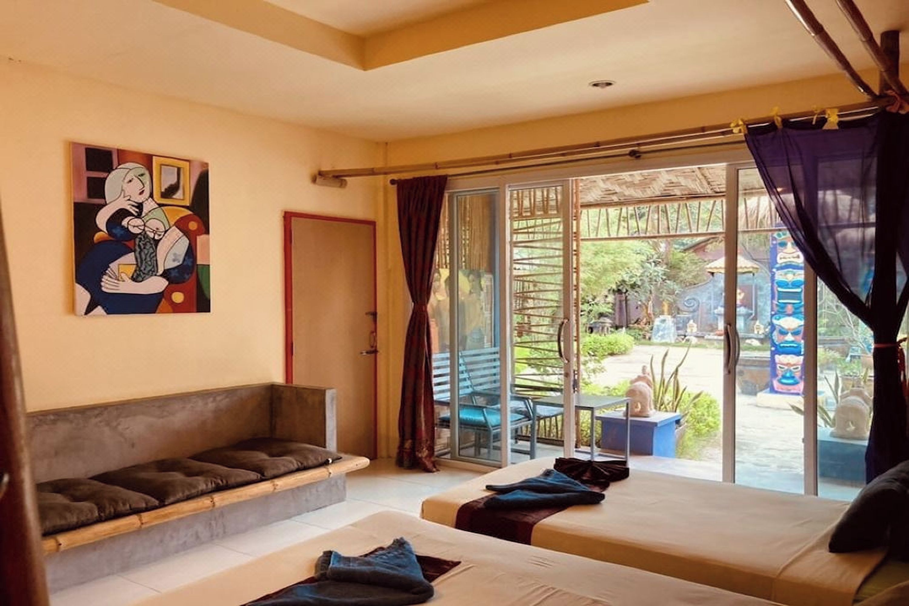 Habitación con dos camas, un sofá y un amplio ventanal corredero que da a un espacio exterior con vistas a zonas verdes y una estatua.