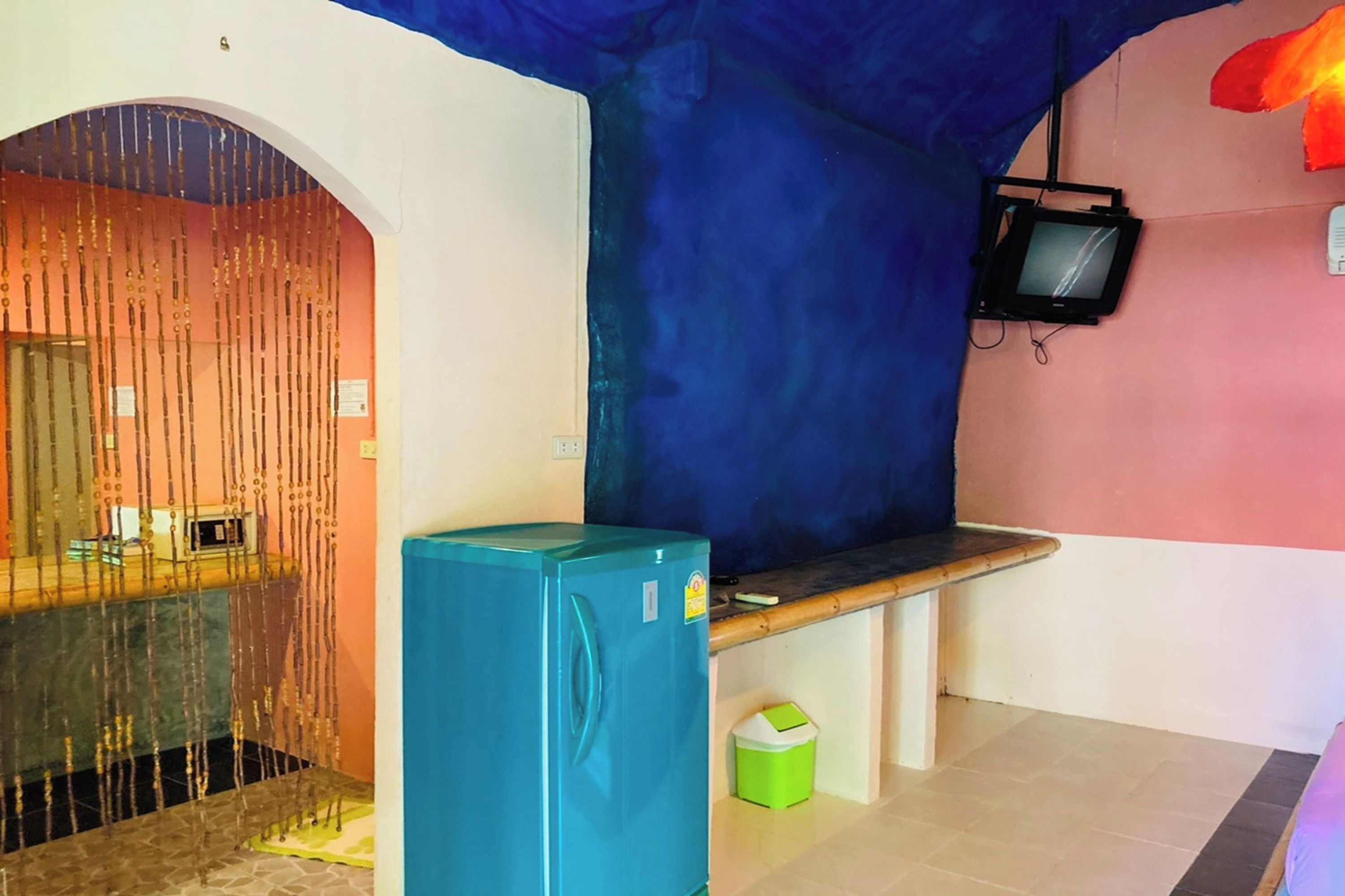 A room with a blue refrigerator, a bench, a television mounted on the wall, and a door with a decorative curtain.