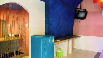 A room with a blue refrigerator, a bench, a television mounted on the wall, and a door with a decorative curtain.