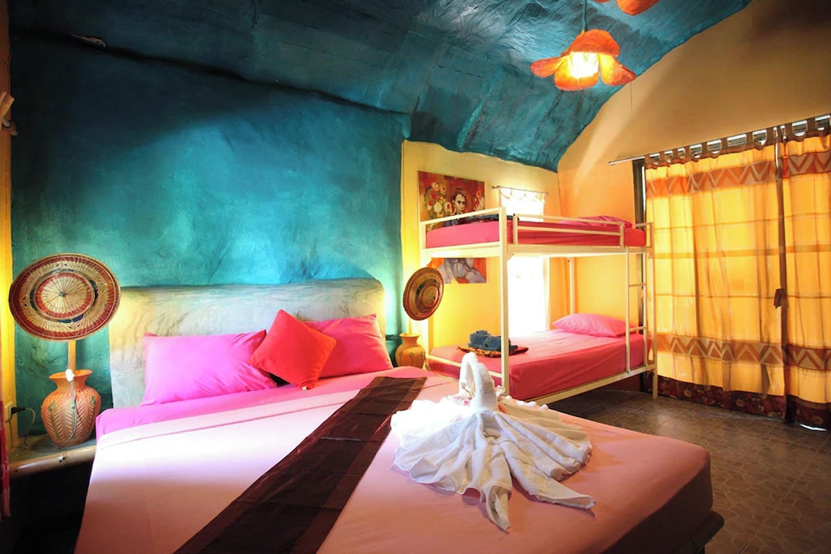 Quarto com beliche, uma cama grande com roupa de cama rosa, um ventilador e um vaso na mesa de cabeceira.