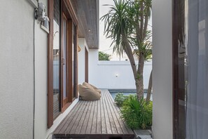 Une petite terrasse en bois, dotée d’une porte-fenêtre coulissante, d’un pouf beige et d’un palmier.