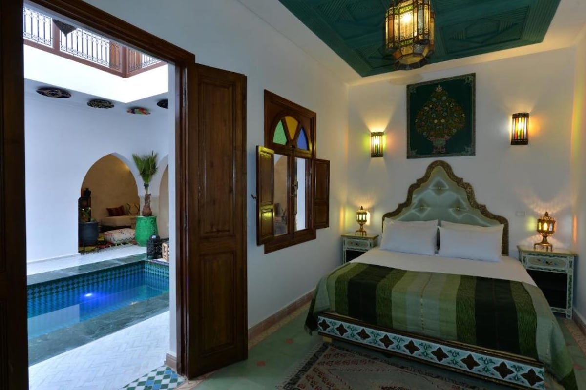 A bedroom with a large bed, a small bedside table, a wall-mounted lamp, and a view of an outdoor swimming pool.