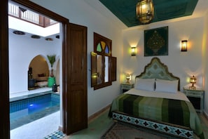 A bedroom with a large bed, a small bedside table, a wall-mounted lamp, and a view of an outdoor swimming pool. - Casa De Marrakech Riad Guest House (Marrakech)