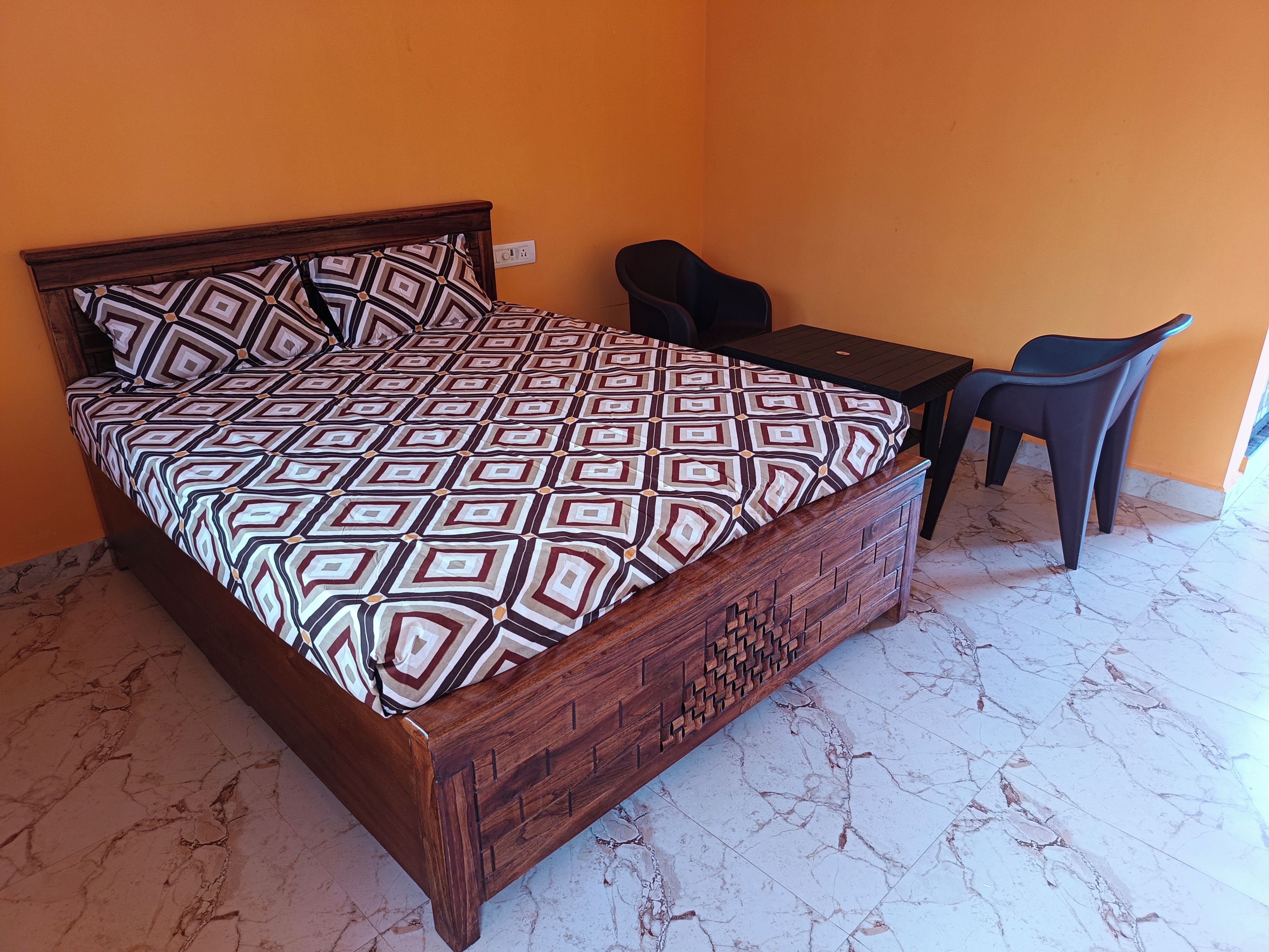 Ein Bett mit geometrisch gemustertem Bettbezug, ein Holz-Kopfteil, ein kleiner Tisch und zwei Stühle in einem Zimmer mit orangefarbenen Wänden und Marmorboden.
