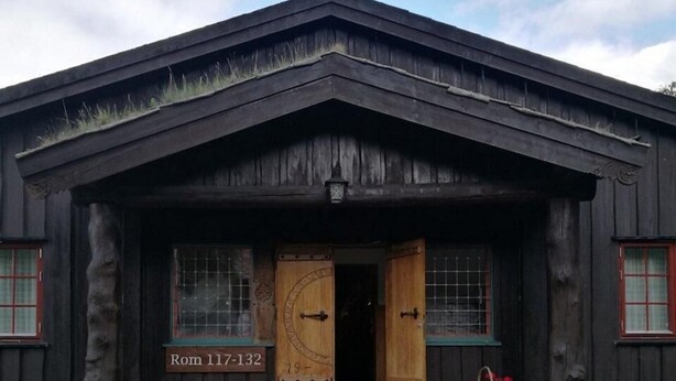 A rustic wooden building with a large wooden door, a small porch, and a sign reading 'Rom 117-132'. - Fossheim Hotell (Lom)