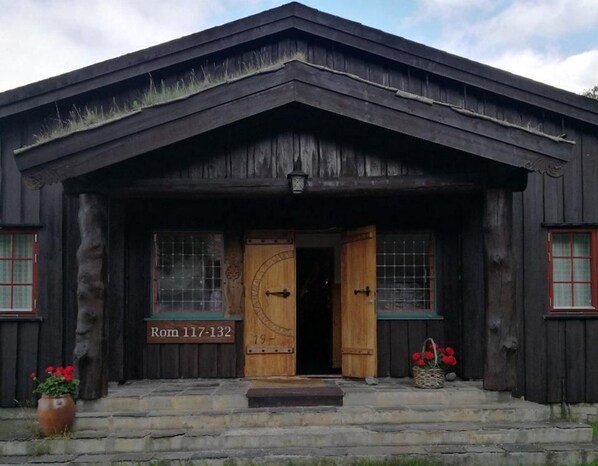 A rustic wooden building with a large wooden door, a small porch, and a sign reading 'Rom 117-132'. - Fossheim Hotell (Lom)