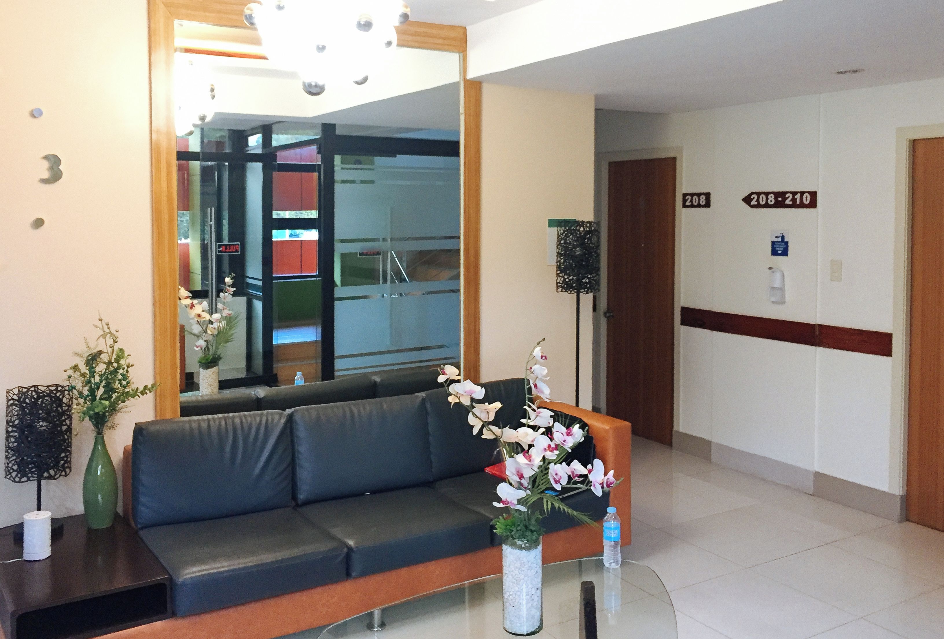 Un área de recepción con un sofá, una mesa de vidrio, un florero y un espejo, además de un cartel que indica las habitaciones 208 al 210.