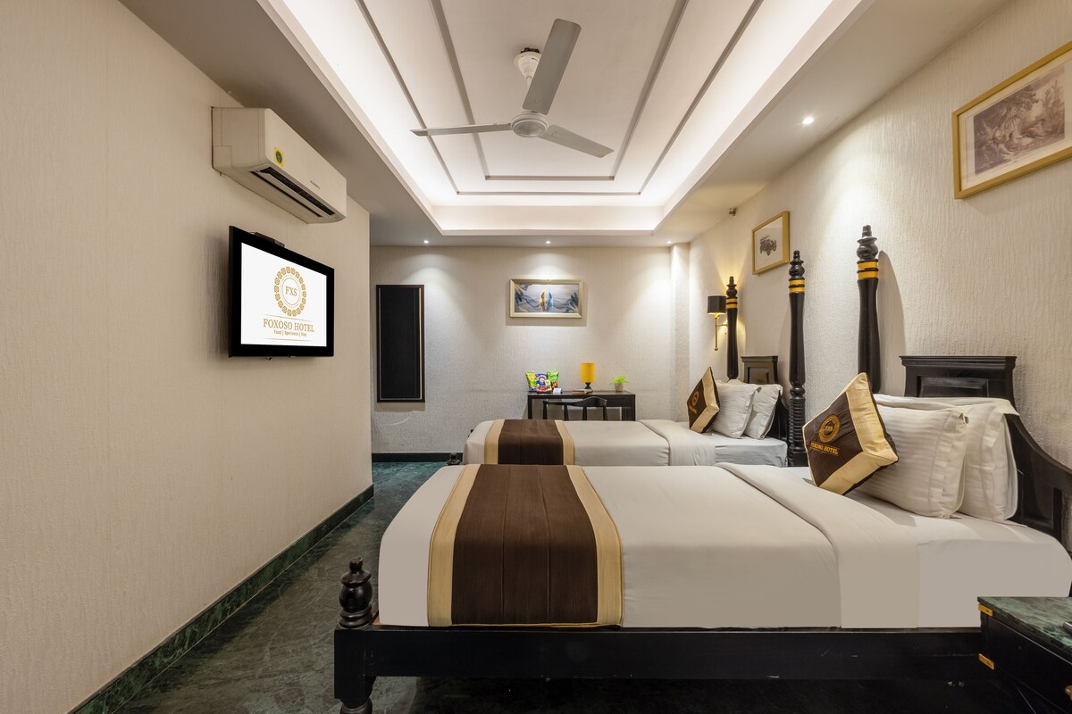 A hotel room with two beds, a ceiling fan, an air conditioning unit, a framed picture on the wall, and a small table with a lamp.