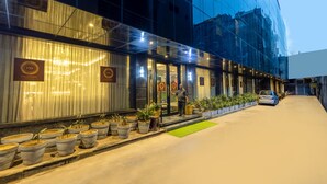 A modern hotel exterior with a glass facade, potted plants, and a car parked in the driveway. - Foxoso Hotel Delhi Airport (New Delhi)