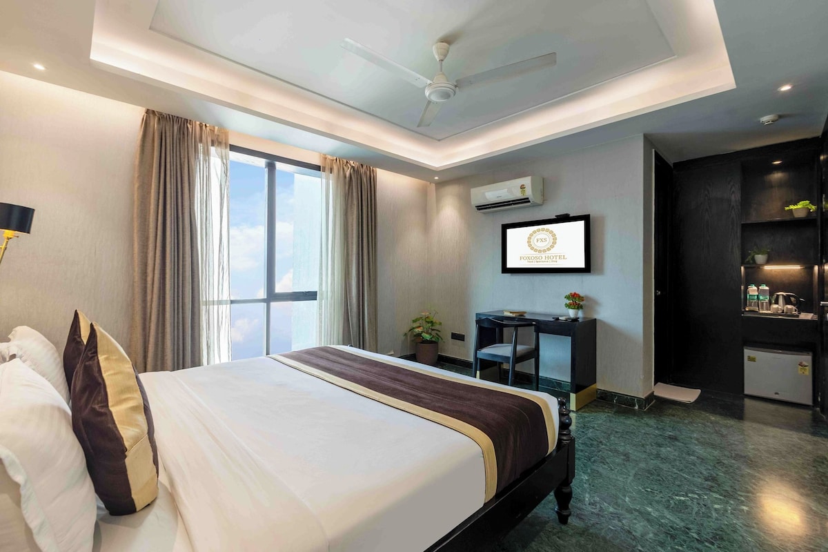 A hotel room with a bed, a desk, a chair, a ceiling fan, an air conditioning unit, and a view of the sky.