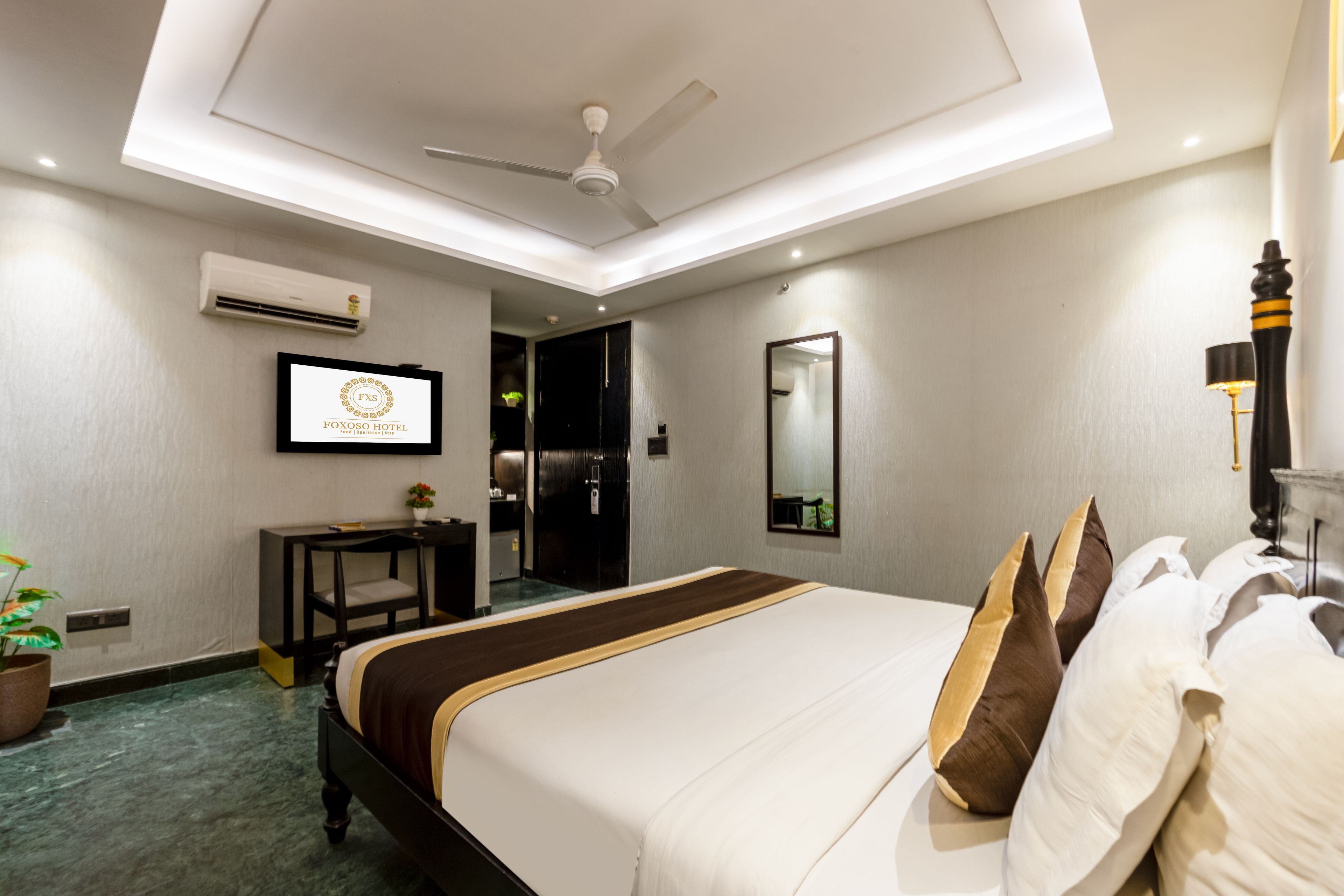 A hotel room with a bed, a ceiling fan, an air conditioning unit, a desk, a mirror, and a television.