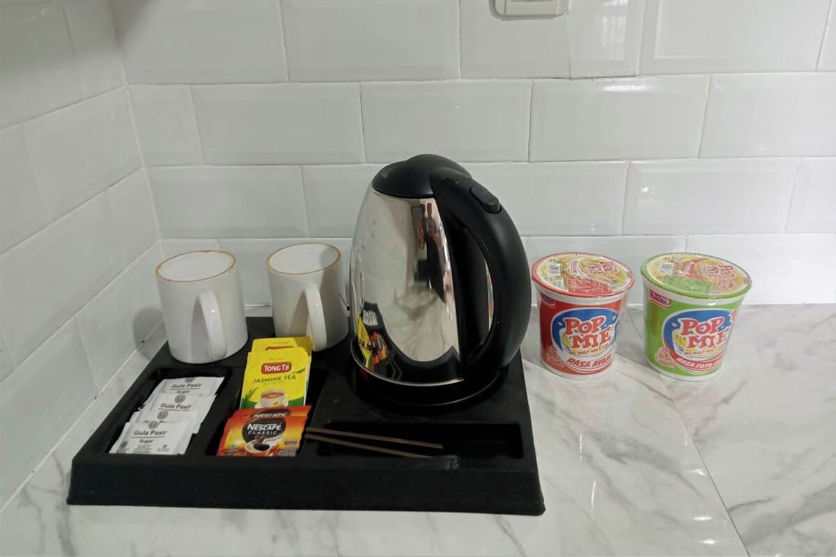 A kitchen counter with a kettle, two mugs, a box of tea, and two containers of instant noodles.