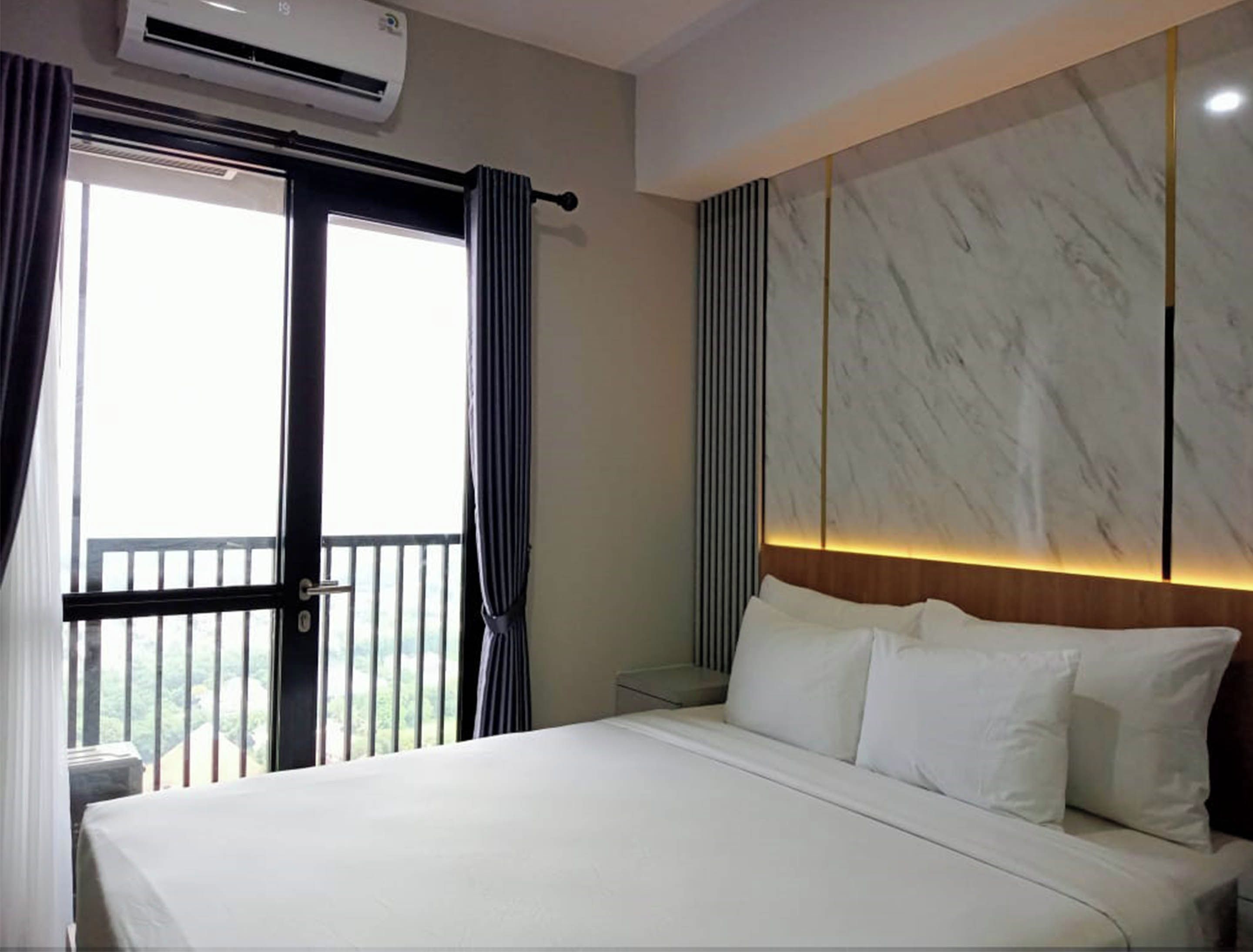 A hotel room with a bed, an air conditioning unit, a window with curtains, and a view of greenery outside.
