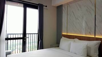 A hotel room with a bed, an air conditioning unit, a window with curtains, and a view of greenery outside.