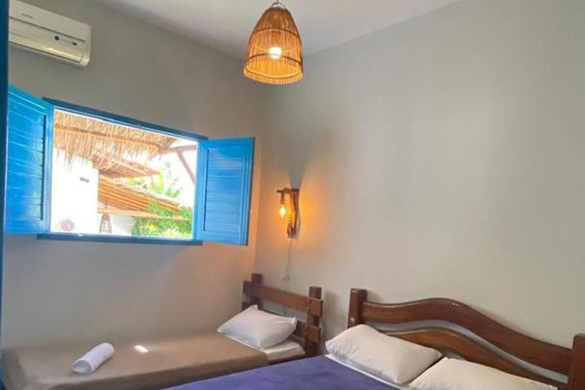 Una habitación con dos camas, una ventana con contraventanas azules, una lámpara colgante y un aire acondicionado instalado en la pared.