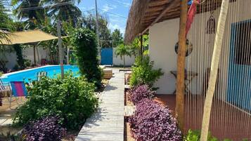 A pool area with a wooden deck, surrounded by lush greenery and a thatched-roof structure.