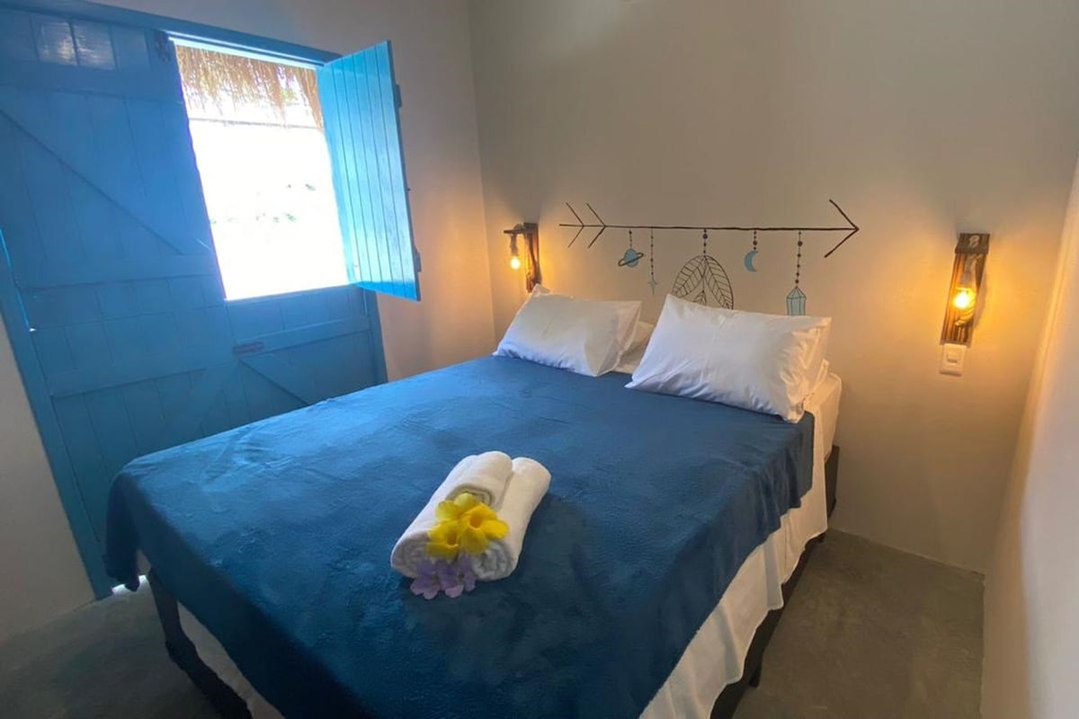 A single bed with a blue bedspread, white pillows, and a towel with flowers. There are two wall-mounted lights and a decorative wall piece above the bed.