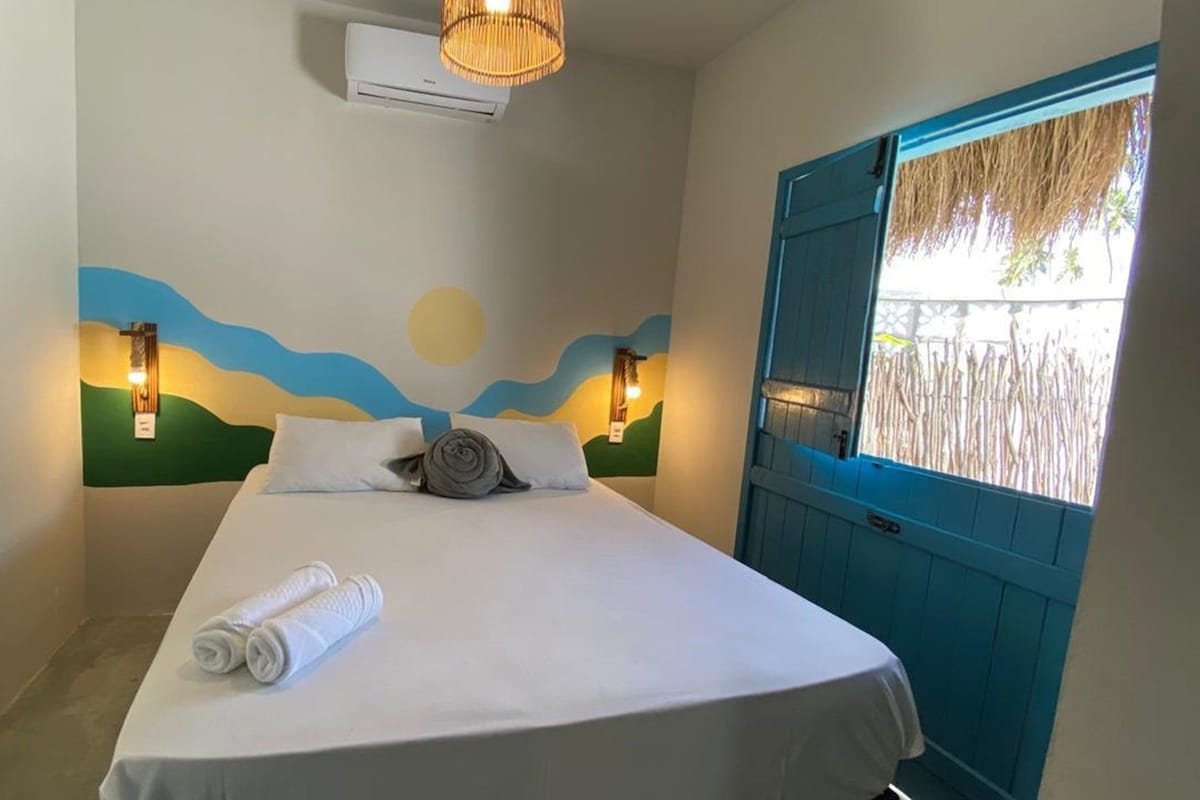 A single bed with white linens, two wall-mounted lights, a ceiling air conditioner, and a window with a blue door.