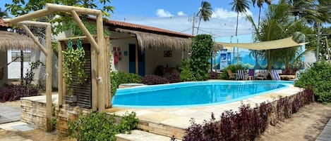 A swimming pool with a wooden pergola, a thatched-roof building, and tropical vegetation.