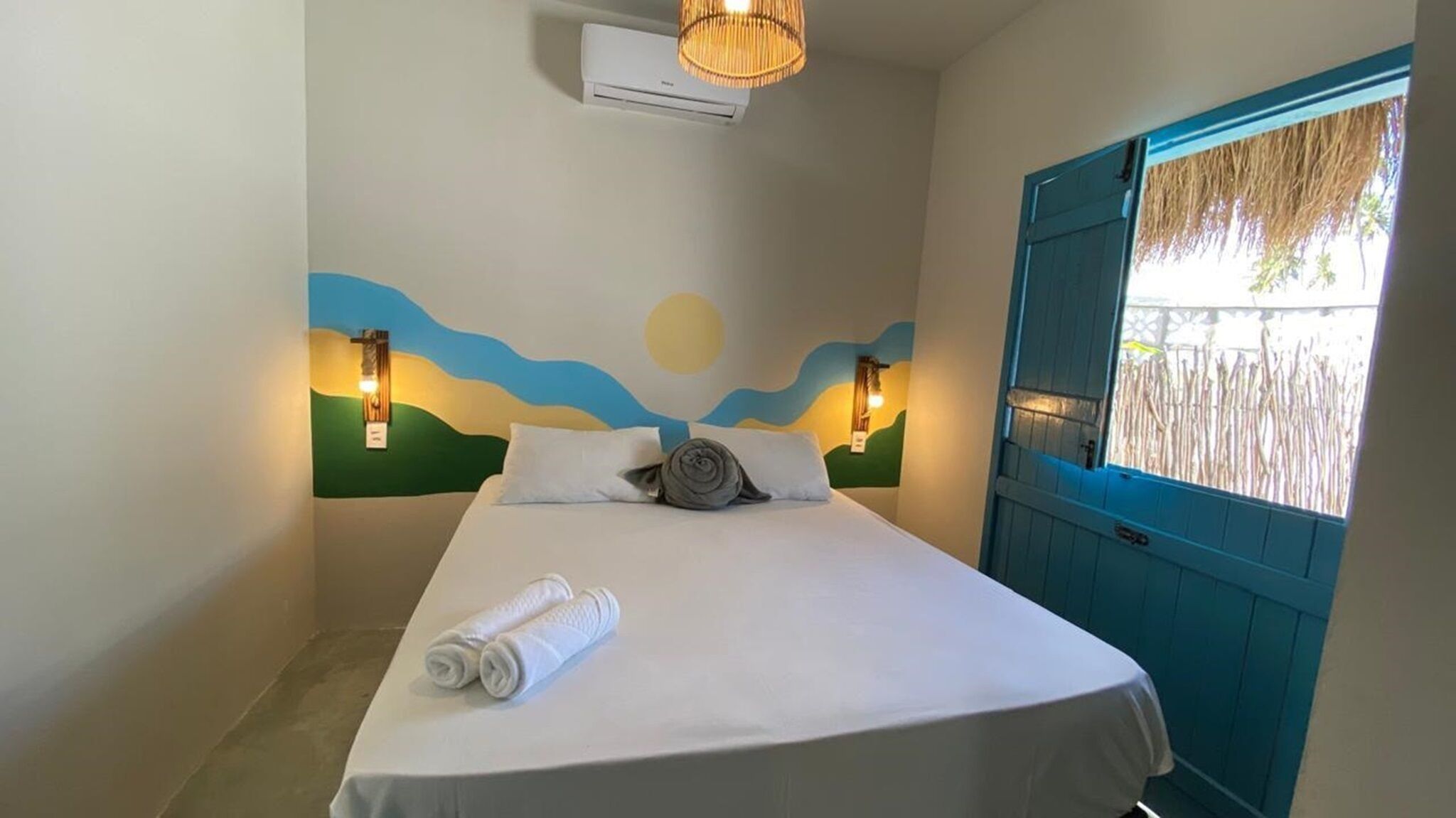 A single bed with white linens, two wall-mounted lights, a ceiling air conditioner, and a window with a blue door.