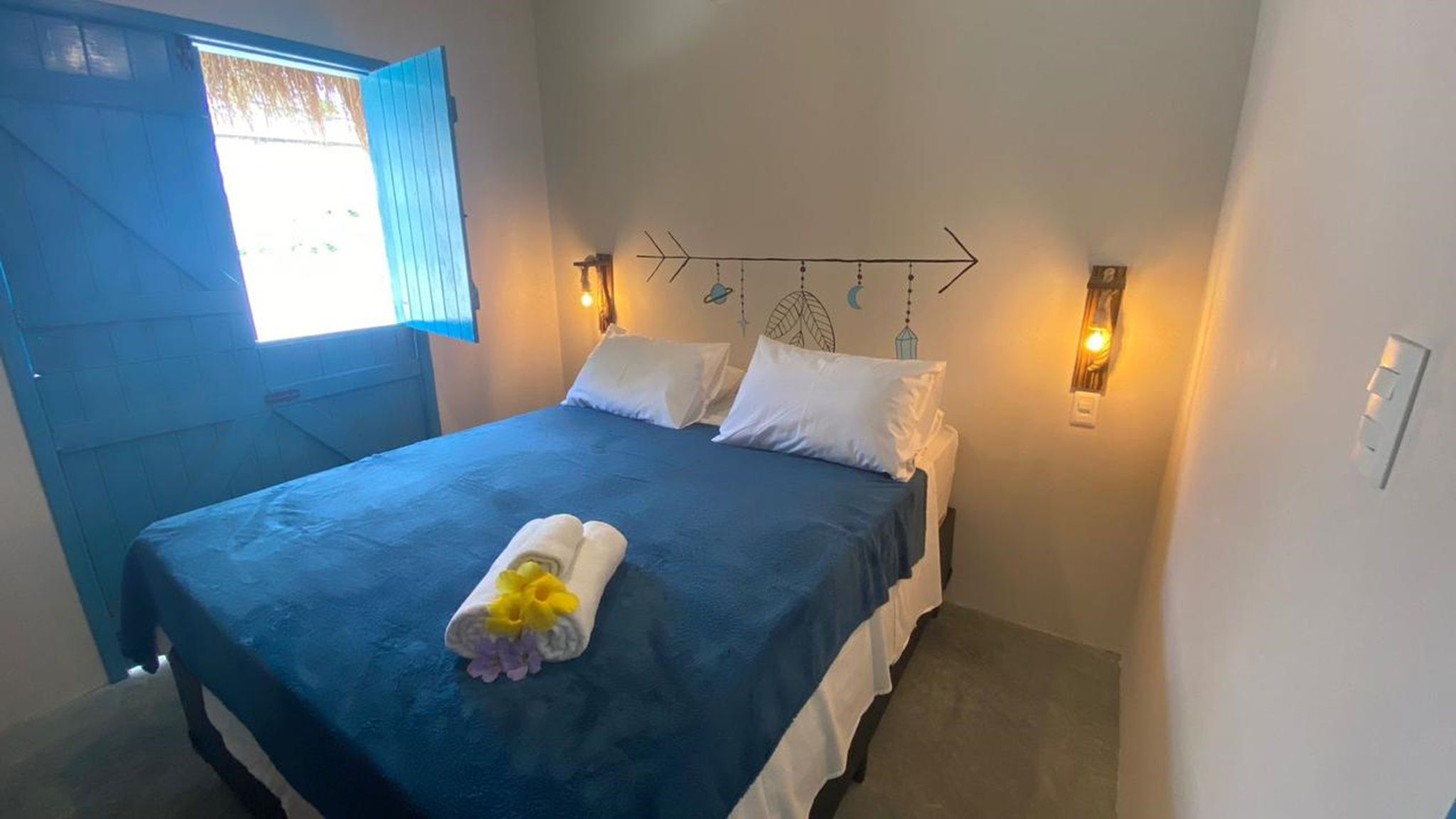 A single bed with a blue bedspread, white pillows, and a towel with flowers. There are two wall-mounted lights and a decorative wall piece above the bed.