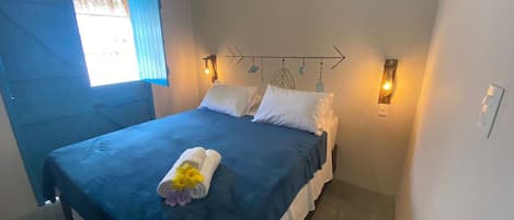 A single bed with a blue bedspread, white pillows, and a towel with flowers. There are two wall-mounted lights and a decorative wall piece above the bed.
