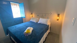 Een enkel bed met een blauwe spreideken, witte kussens en een handdoek met bloemen. Er zijn twee wandlampen en een decoratief object aan de muur boven het bed.
