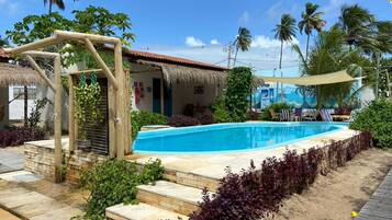 A swimming pool with a wooden deck, a thatched-roof structure, and tropical vegetation.