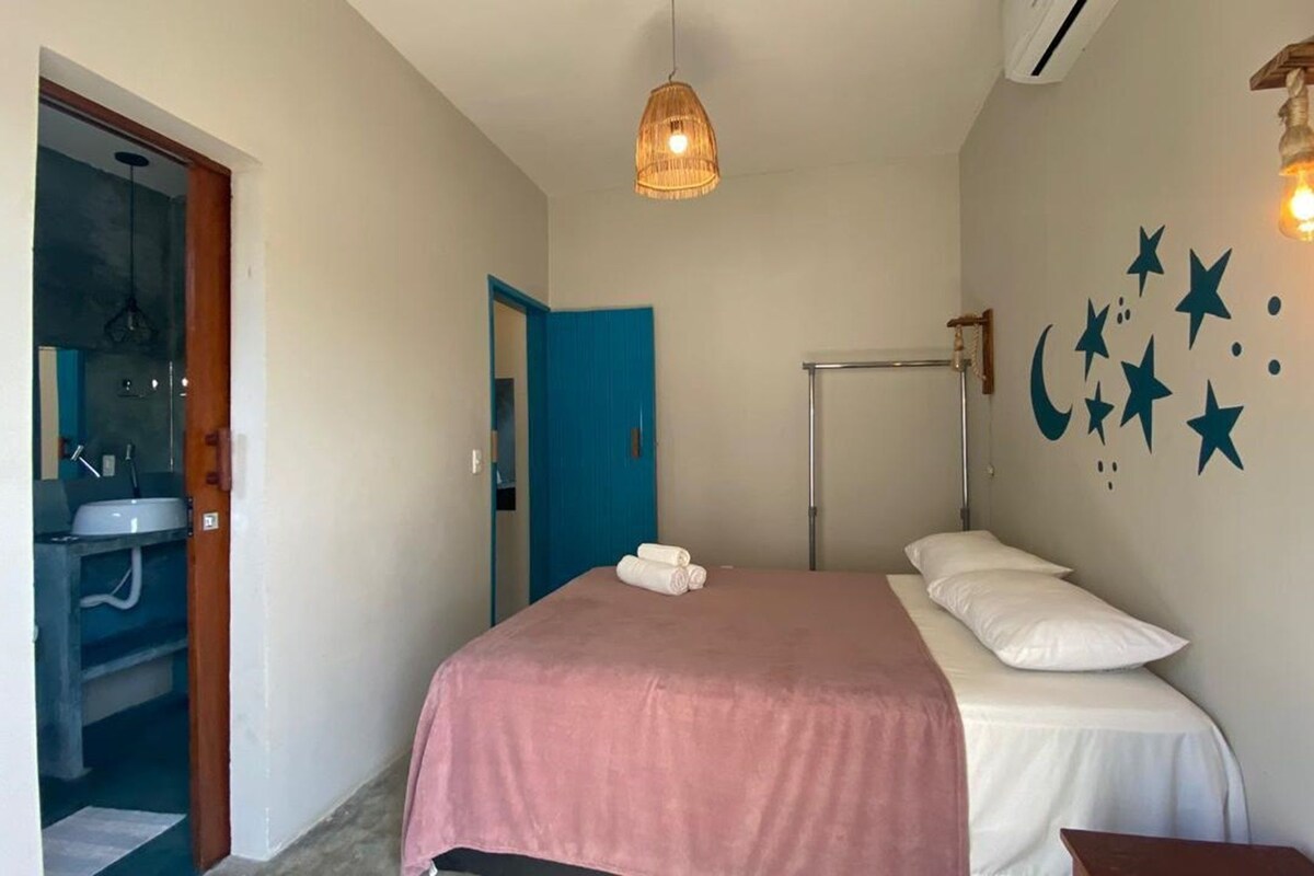Un dormitorio con cama, mesita de noche, lámpara colgante, aire acondicionado instalado en la pared y un baño visible a través de una puerta abierta.