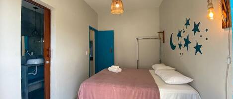 A bedroom with a bed, a bedside table, a hanging light fixture, a wall-mounted air conditioner, and a bathroom visible through an open door.