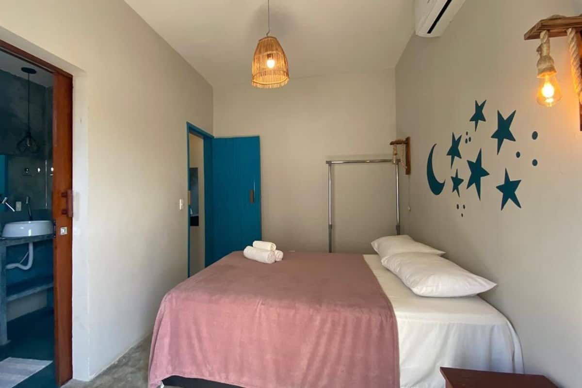 Un dormitorio con cama, mesita de noche, lámpara colgante, aire acondicionado instalado en la pared y un baño visible a través de una puerta abierta.