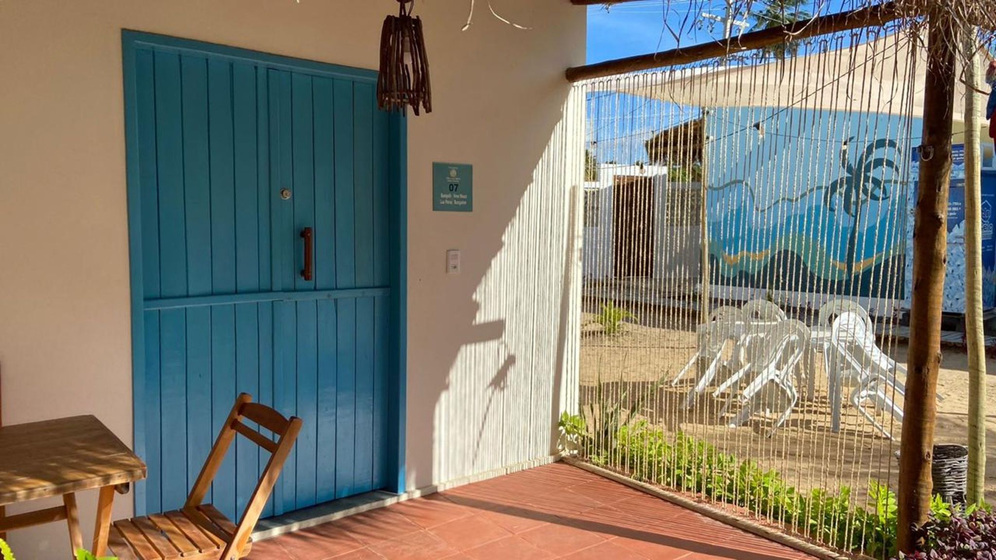 Una habitación con una puerta azul, una silla, una mesita y vistas a través de una ventana con cortina blanca.