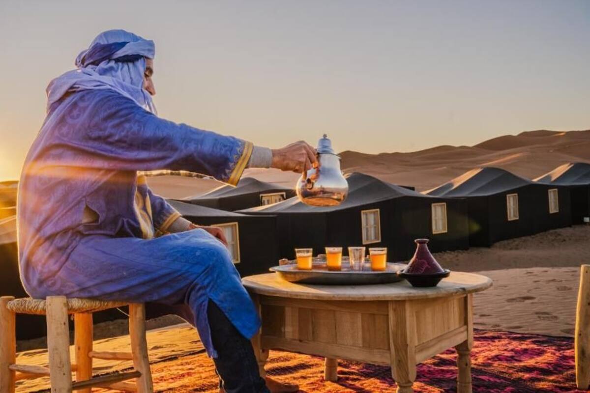 A person sitting at a wooden table with a tray of drinks and a teapot, overlooking tents in a desert setting.