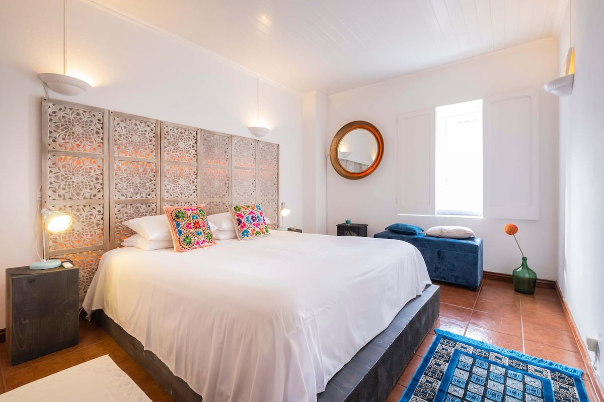 Un dormitorio con una cama grande, una mesita de noche con lámpara, un cabecero decorativo, un espejo redondo, un sofá azul y un jarrón con una flor.