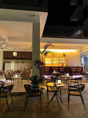 A well-lit outdoor seating area with tables and chairs, a bar area with shelves of bottles, and a large plant in the center. - Angel Garden Resort (Busuanga)